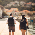 Two people with backpacks walking along a coastal street.